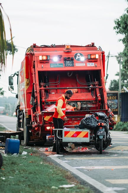 A worker in a yellow and red uniform operates a large red rubbish collection vehicle on a roadway, with the rear loading mechanism open and partially filled with waste debris. The vehicle is positione