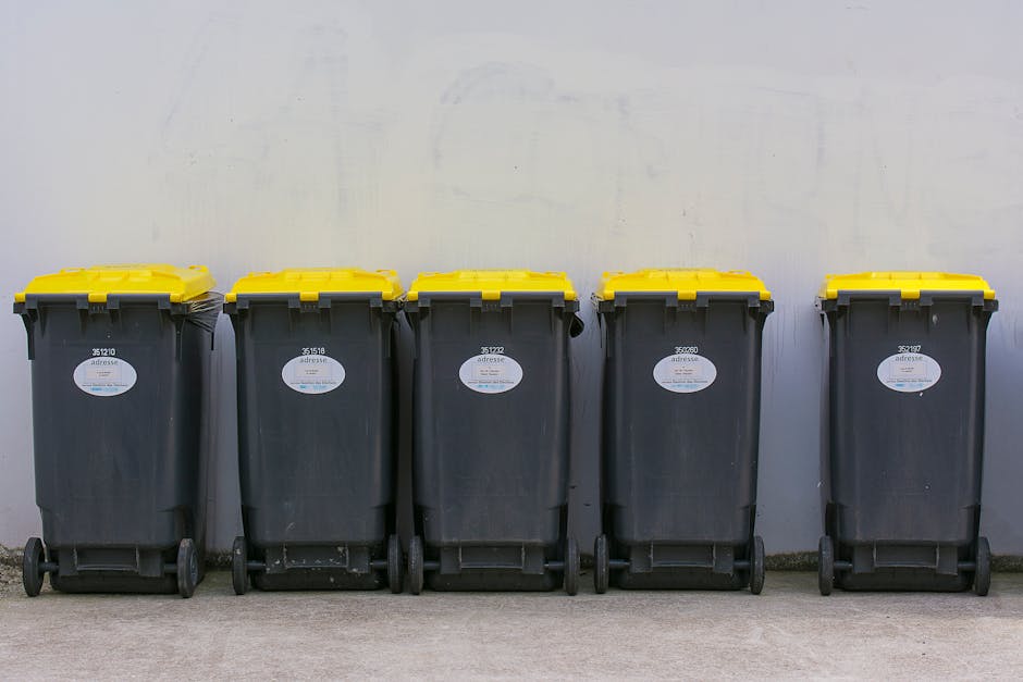 Five large wheeled rubbish bins made of dark grey plastic are lined up along a concrete surface against a plain, light-colored wall. Each bin features a textured surface with vertical ridges and is eq