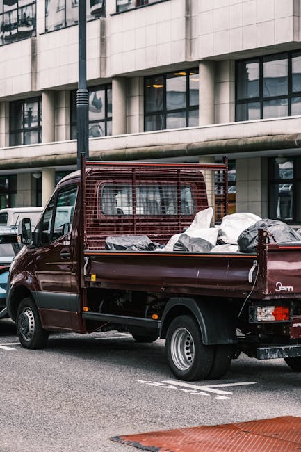 The image displays the rear side of a white commercial van parked on a paved street, with three large blue rubbish bags placed on the ground against the back of the vehicle. The bags appear to be fill